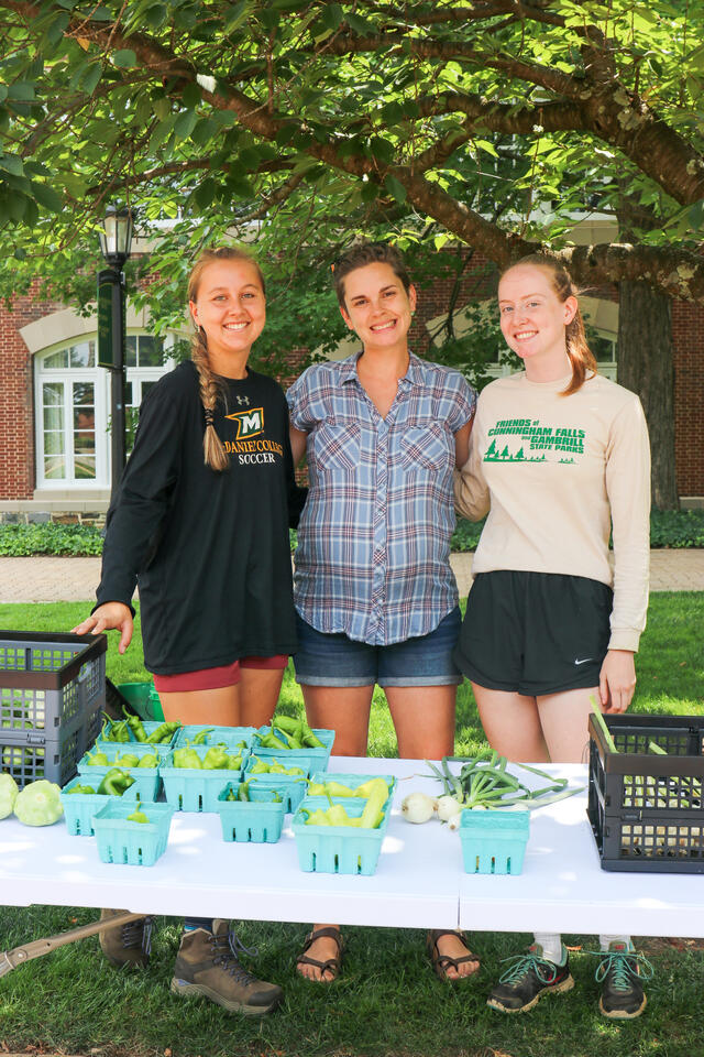 Professor Elly Engle poses with two students behind a table with produce on it.