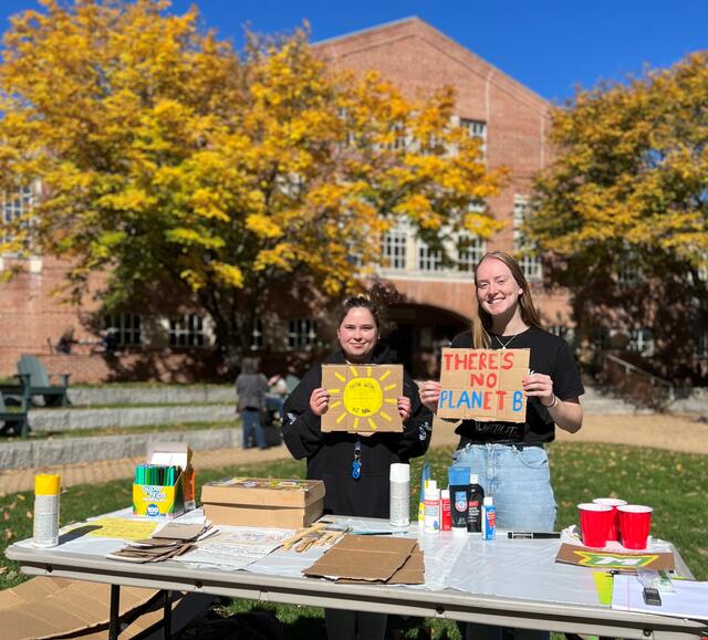 Two students hold up handmade signs at Global Climate Change Week. One shows a sun and the other reads "There's no planet B."