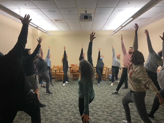 People stand in circle with extended arms in a bare room 