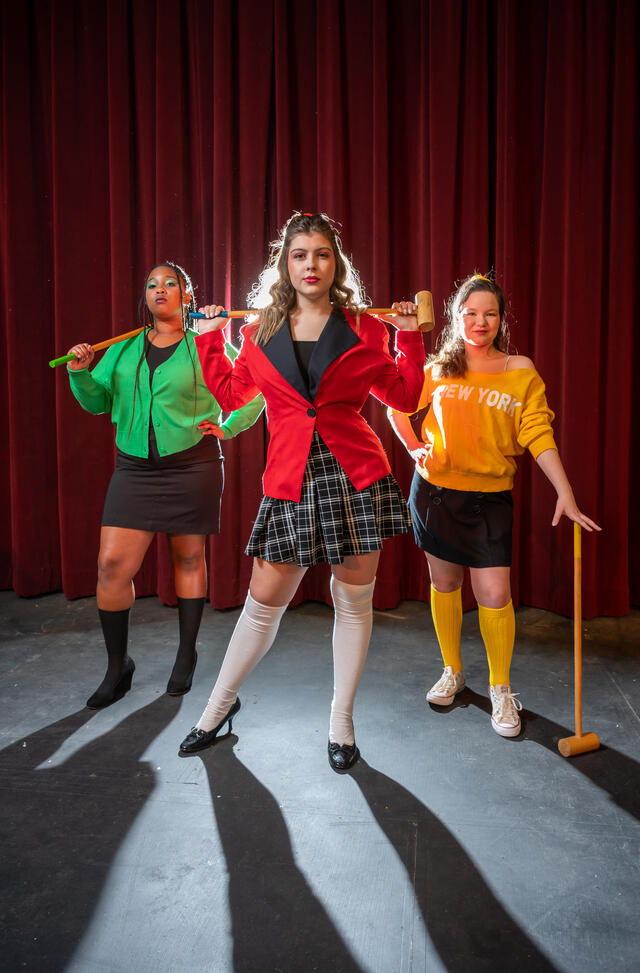 Three students posing in vivid colored clothing in front of a red curtain with mallets. 