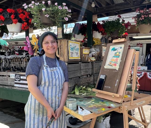 Sarah Mendez at an easel wearing an apron. Behind her is a market stand with flowers.