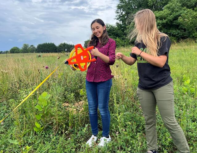 Two students stand in a field, one holding the end of a large tape measure while the other shows her how to use it.