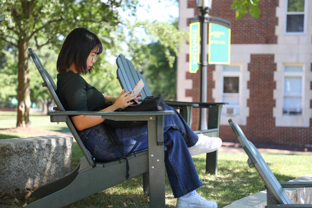 A students sits and reads in a green Adirondack chair on campus.