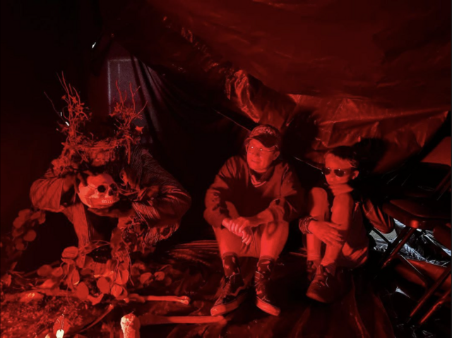 Steven Angle sits with a young boy in a tent lit with red light next to someone in a costume holding a skull.