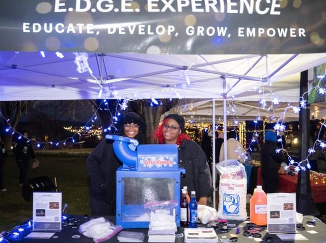 Two students stand behind a sno cone ice machine at a table with a banner above them reading EDGE Experience.