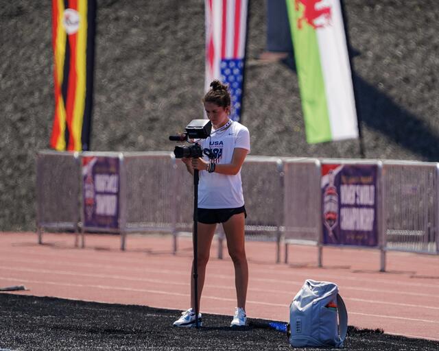 A woman stands holding a camera looking out of frame. Flags for USA, Wales, and Germany are behind her.