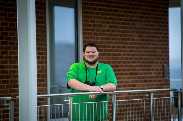 A student leans forward on a railing with a pair of binoculars around his neck.
