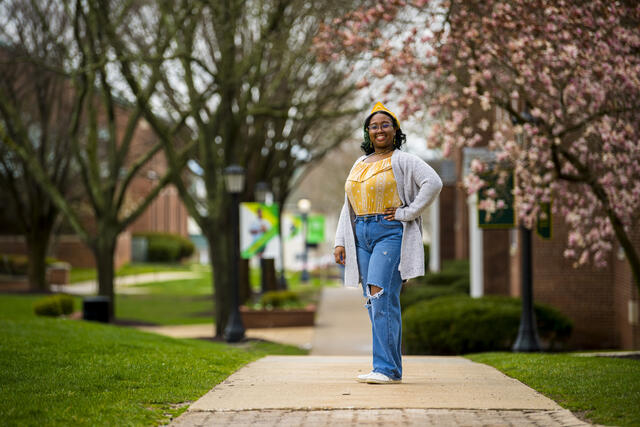 A student in a yellow shirt and grey cardigan stands outside in front of a row of cherry blossom trees.