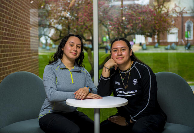 A pair of twin sisters sits at a table in front of a window.