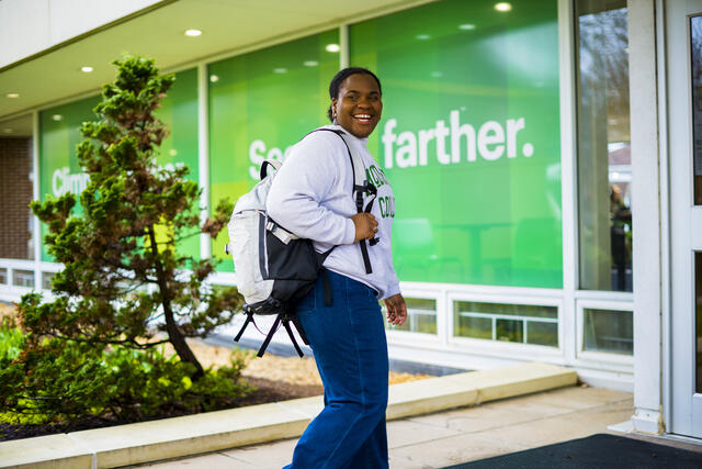 A student smiles to the side while walking with a backpack on their shoulder.