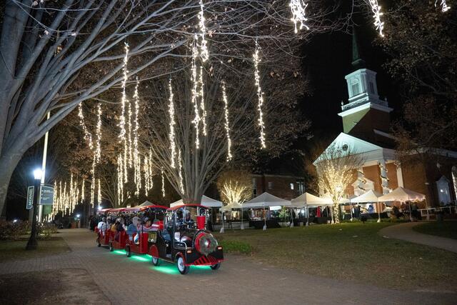 A small train car with passengers drives under trees with strings of white lights in them.