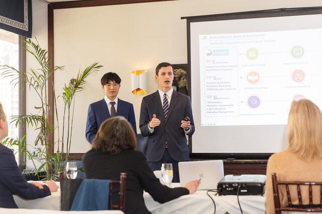 A white man in a suit stands and presents in front of a screen at a conference. Another man in a suit stands beside him.