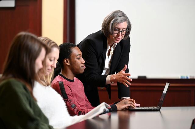 A woman in a black blazer leans over a row of seated students with laptops.