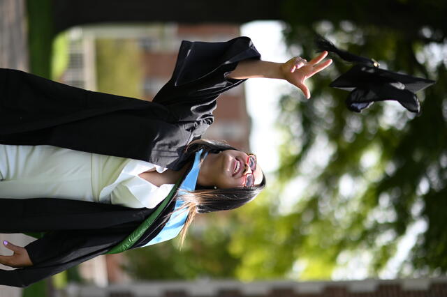 A female student in a white dress, glasses, and commencement regalia tosses her graduation cap in the air.