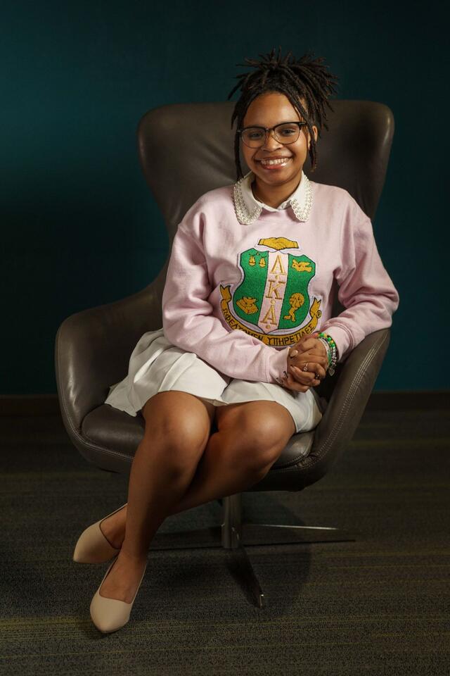 Bria Hill sits in an armchair. She wears a white skirt, white shoes, and a pink sweatshirt with the logo for Alpha Kappa Alpha Sorority.
