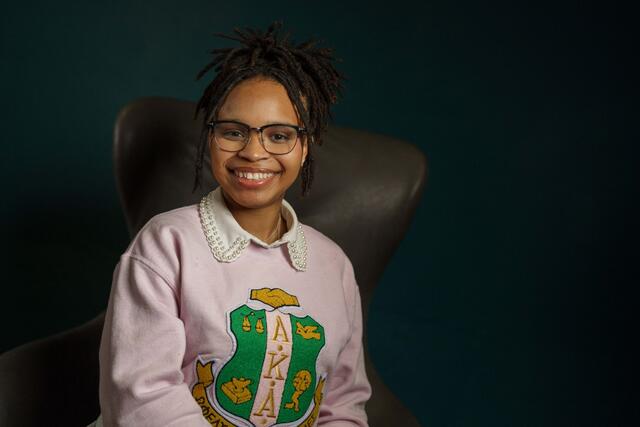 Bria Hill sits in an armchair. She wears a pink sweatshirt with the logo for Alpha Kappa Alpha Sorority.