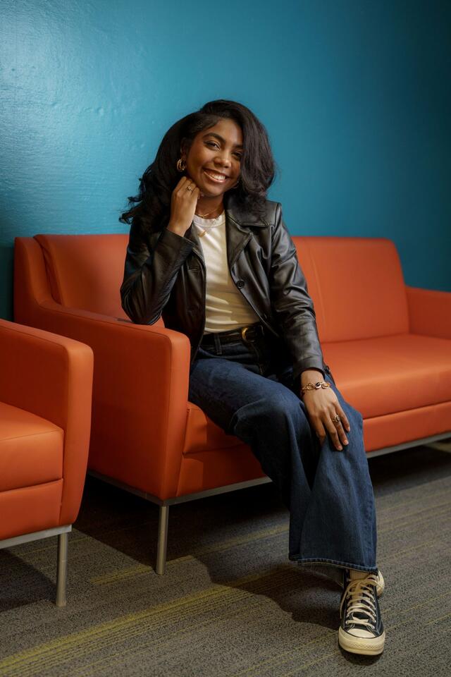 Portrait of Janiah Fields sitting on an orange couch in front of a blue wall, wearing jeans, a white shirt, and a black leather blazer.