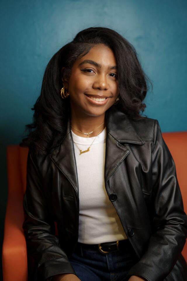 Portrait of Janiah Fields sitting on an orange couch in front of a blue wall, wearing a white shirt and a black leather blazer.
