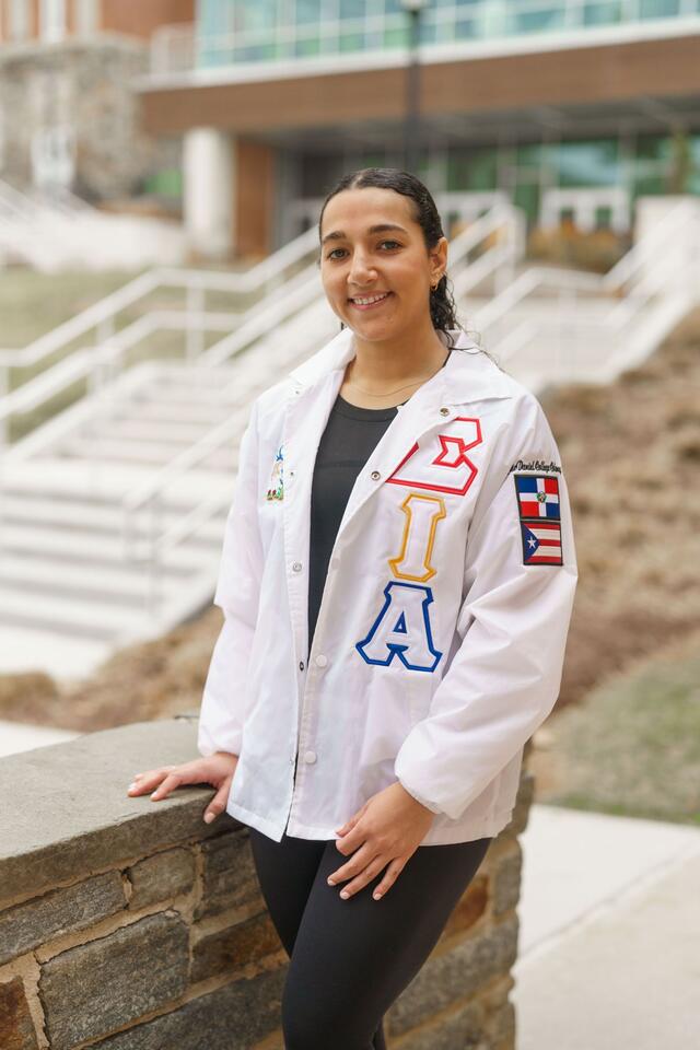 Katherine Taveras poses outside of the student center. She wears a white jacket with the logo for her sorority, Hermandad de Sigma Iota Alpha.