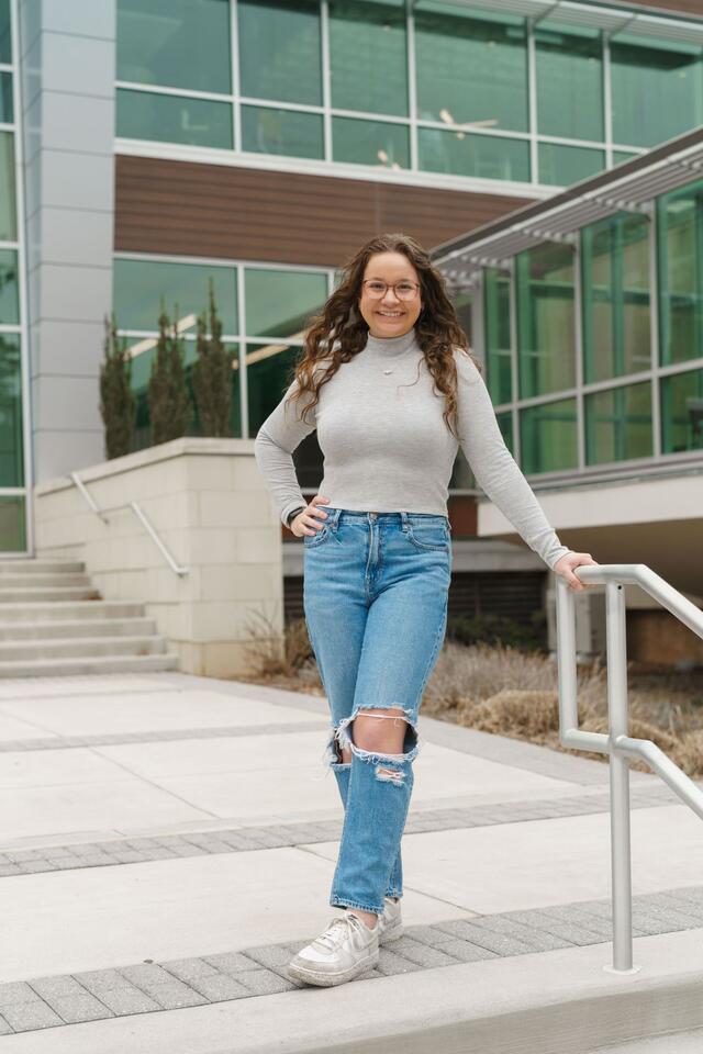 Mackenzie McCarter stands outside the student center wearing a grey long-sleeved shirt and jeans.