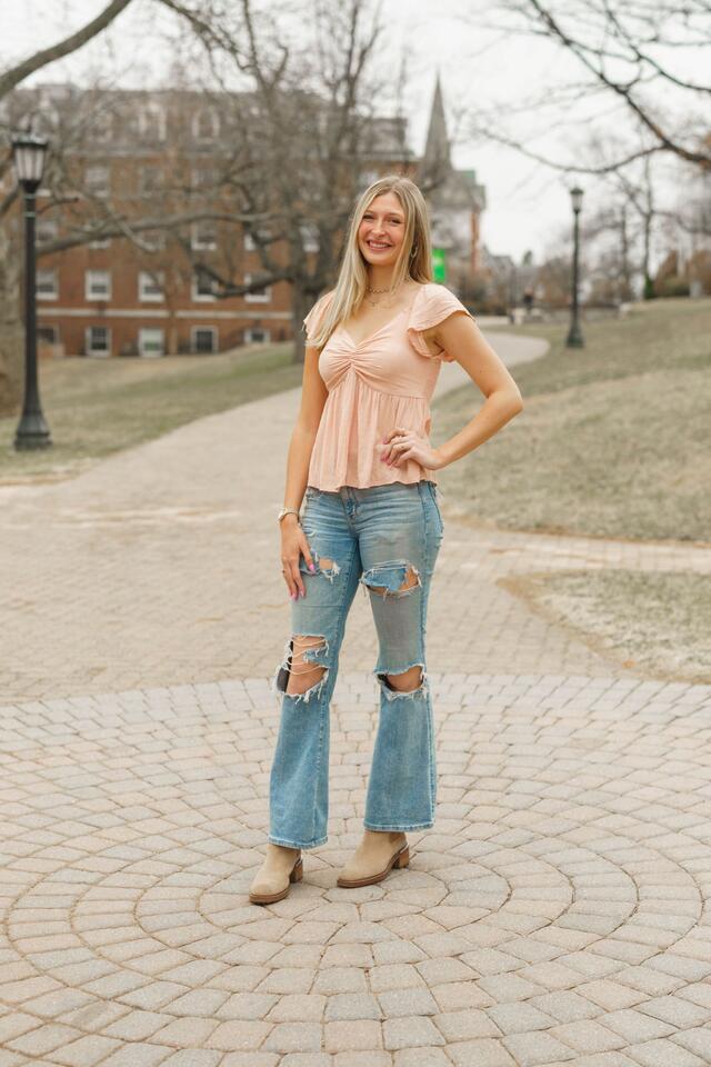Madge Myers stands outside with one hand on her hip, wearing a pink blouse and jeans.