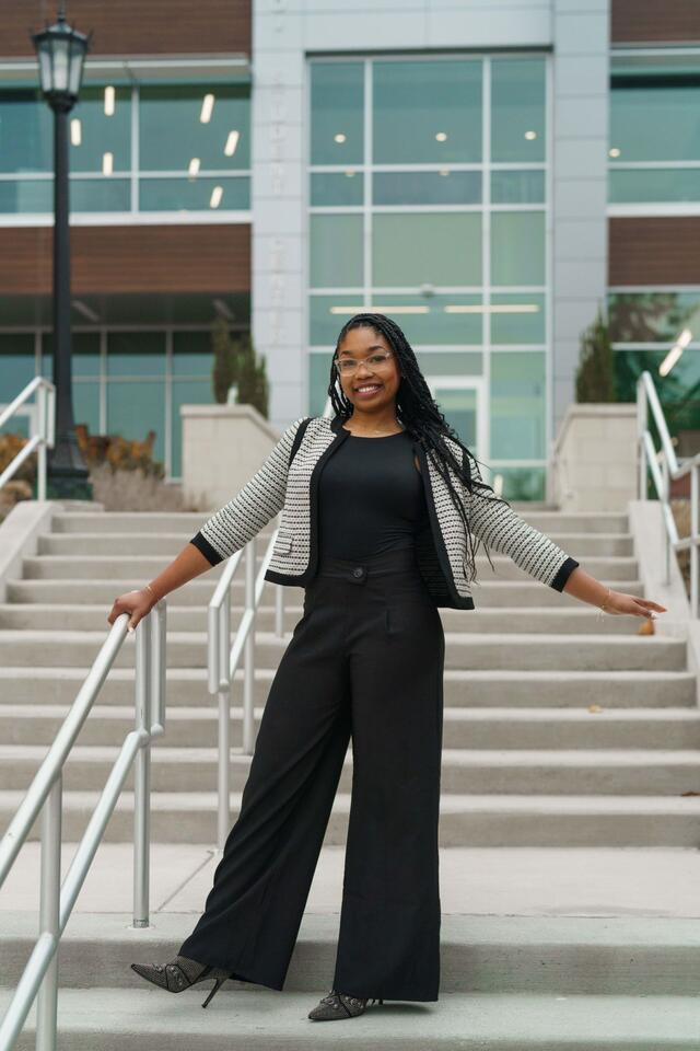 Nakayla Lawson stands outside of the student center wearing a black shirt and pants with a black and white blazer. She has one hand on a railing and both hands held out to her sides.