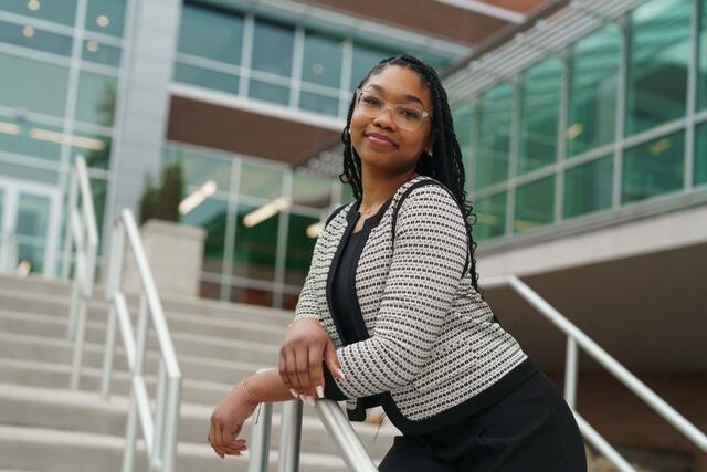 Nakayla Lawson stands outside of the student center wearing a black shirt and pants with a black and white blazer. She leans forward on a railing.