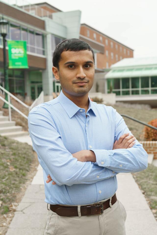 Nikhil Niyogi stands outside of the student center with his arms crossed, wearing a blue button-down shirt, a belt, and tan pants.