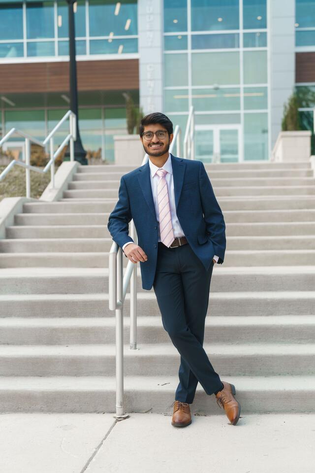 Parth Shah stands outside in front of a set of stairs wearing a navy suit and pink striped tie.