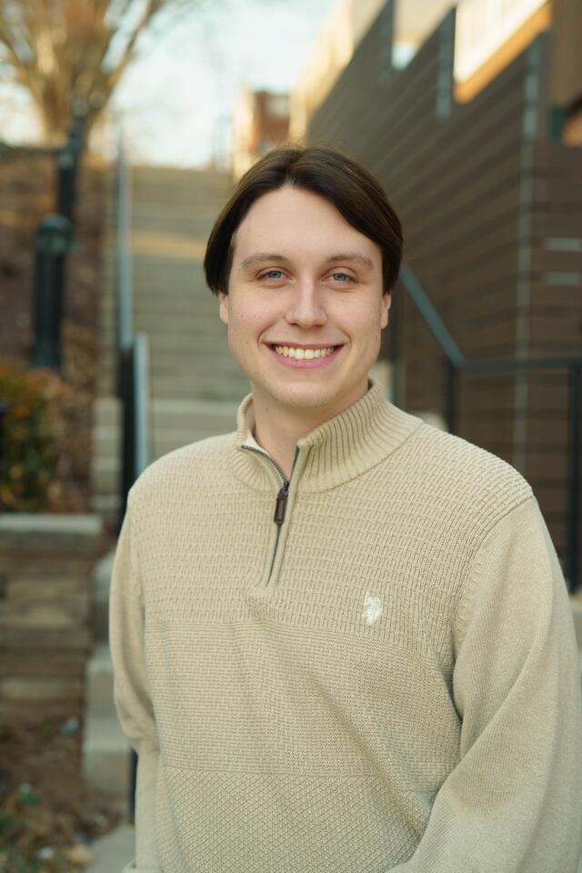 Headshot of Paul Steinert standing outside in front of a staircase, wearing a tan sweater.