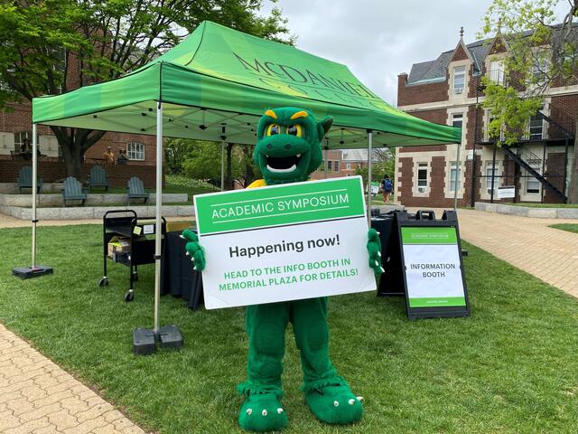 The Green Terror mascot holds a sign for the Academic Symposium while standing outside in front of a green tent.