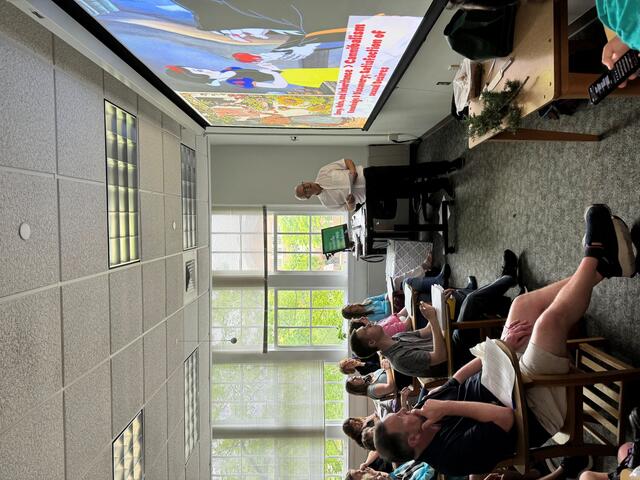 A man in a white shirt stands at the front of a classroom in front of a projector screen, while an audience listens.