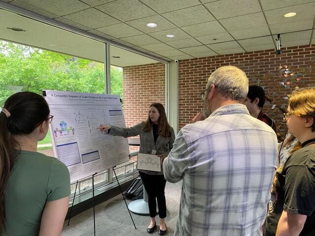 A student points to a poster board featuring research while a group listens.