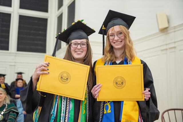 2 students in graduation regalia pose with gold award envelopes
