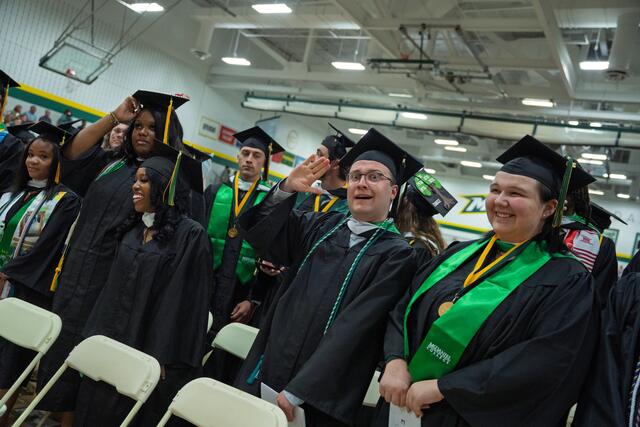 students in graduation regalia stand behind seats
