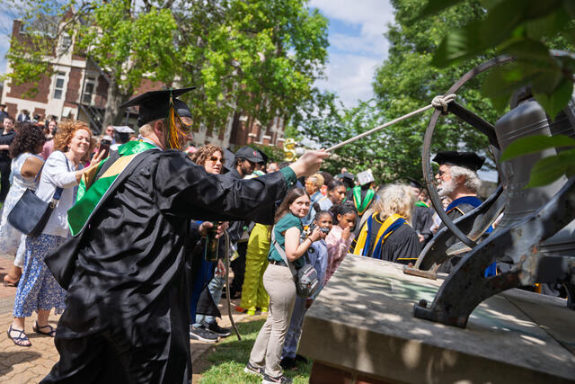Student in graduation regalia pulls on string to ring large bell with a crowd in the background