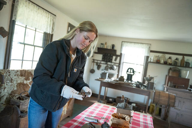 Alicia Heard wears white gloves while standing in a historical kitchen and handling heirloom kitchen utensils.
