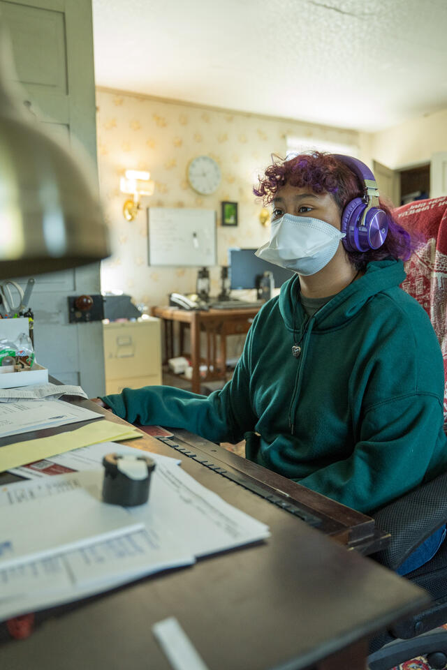 A studet in purple headphones and green sweatshirt sits at a computer desk in the homestead office.
