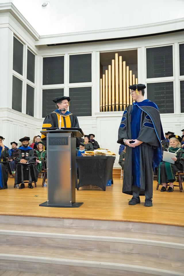Man wearing regalia stands at podium looking toward another man in regalia