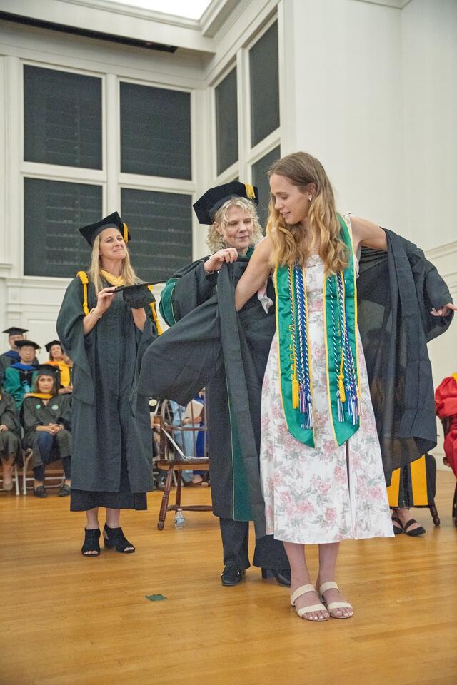 student wearing graduation cords is helped into a robe by president Jasken, with faculty looking on