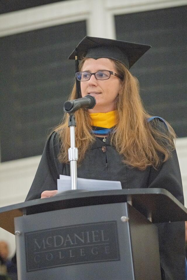 Woman wearing graduation regalia speaks at a podium with a McDaniel College sign on the front