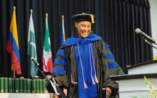 Woman in graduation regalia stands on stage smiling with flags in a row behind her
