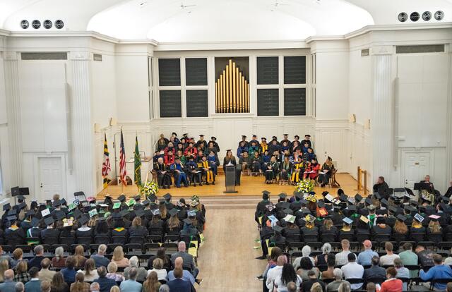 Rear view of audience in chairs, including students wearing graduation regalia, facing a stage with a woman wearing regalia speaking at a podium with people seated in rows behind her also wearing regalia