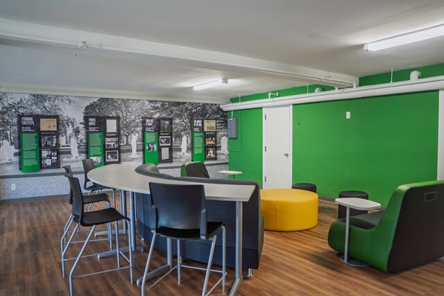View of the clubroom with a curved high-top table and chairs facing 2 walls - one green and the other with wall displays