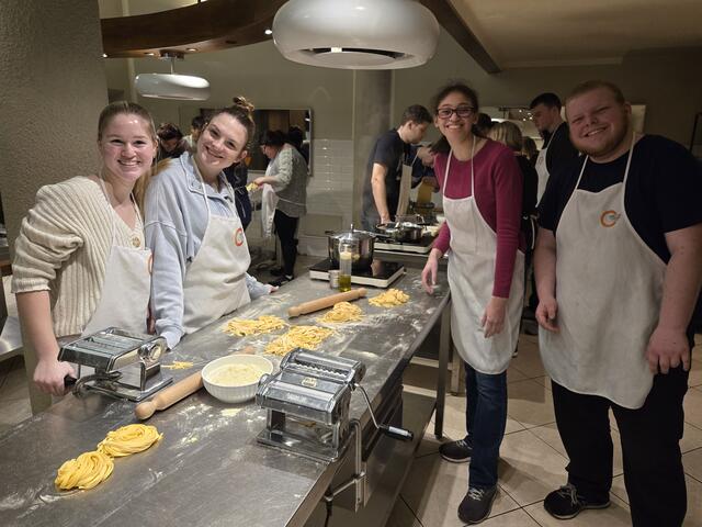 A group of students in white aprons stand in a kitchen for a cooking class making pasta.