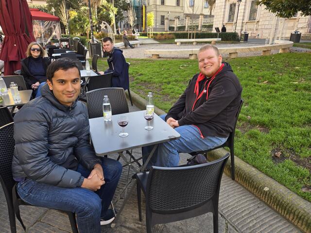 Two students sit together at a table outside with drinks in front of them.