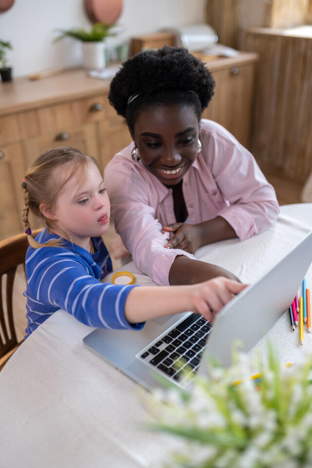 A young woman points to a laptop with a young girl who has special needs.