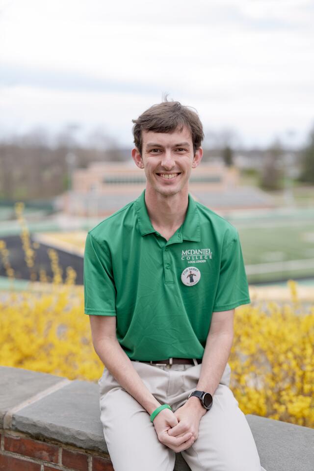 A male student in a McDaniel polo shirt sits in front of the football field.