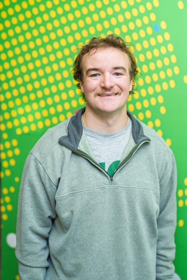 A male student smiles in front of a green patterned wall.