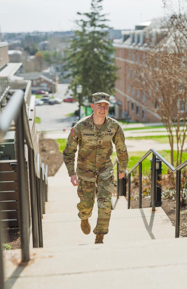 A male student in a military uniform walks up the steps outside of Roj Student Center.